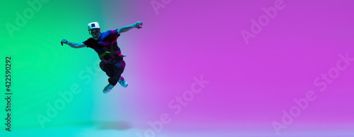 Beautiful sportive boy dancing hip-hop in stylish clothes on colorful gradient background at dance hall in neon light.