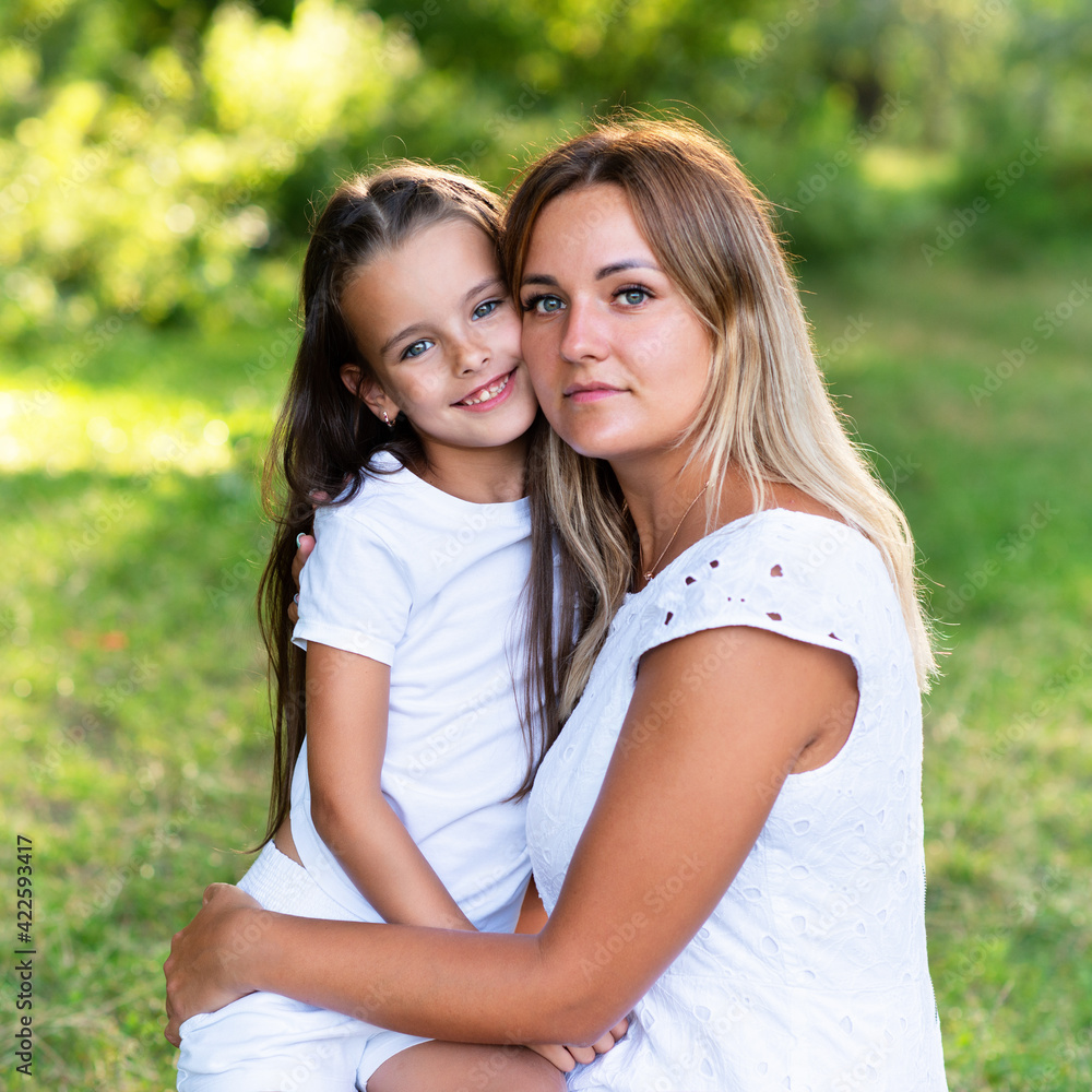 Little girl hugs her mother in summer forest nature outdoor. Portrait of mom and daughter wearing white clothes against summer greenery. Little girl hugs her mother in summer forest nature outdoor. Portrait of mom and daughter wearing white clothes against summer greenery.