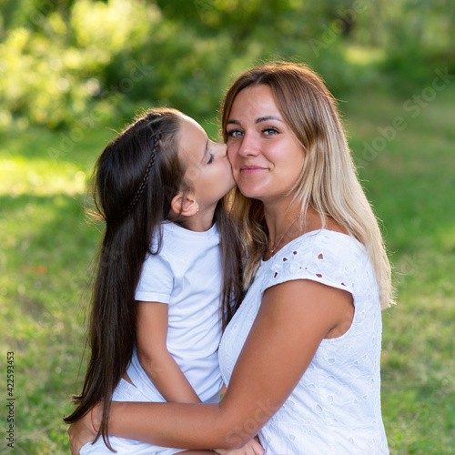 Little girl hugs her mother in summer forest nature outdoor. Portrait of mom and daughter wearing white clothes against summer greenery. 