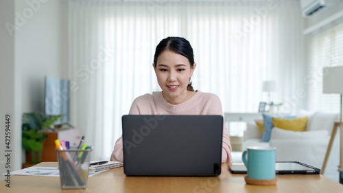 Candid young attractive beautiful asian businesswoman work remotely from home or learning online sitting smiling in cozy workspace at home feeling happy in quarantine social distance concept.