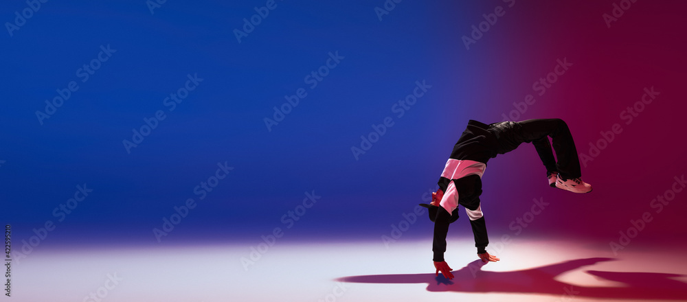 Beautiful sportive boy dancing hip-hop in stylish clothes on colorful gradient background at dance hall in neon light.