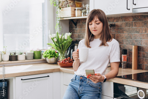 Close up woman in white t-shirt drinking coffee or tea and using phone in kitchen at home. Early breakfast in the morning, rest time, break from work