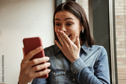Φωτογραφία amazed woman in wireless earphones listening music and covering mouth while hold