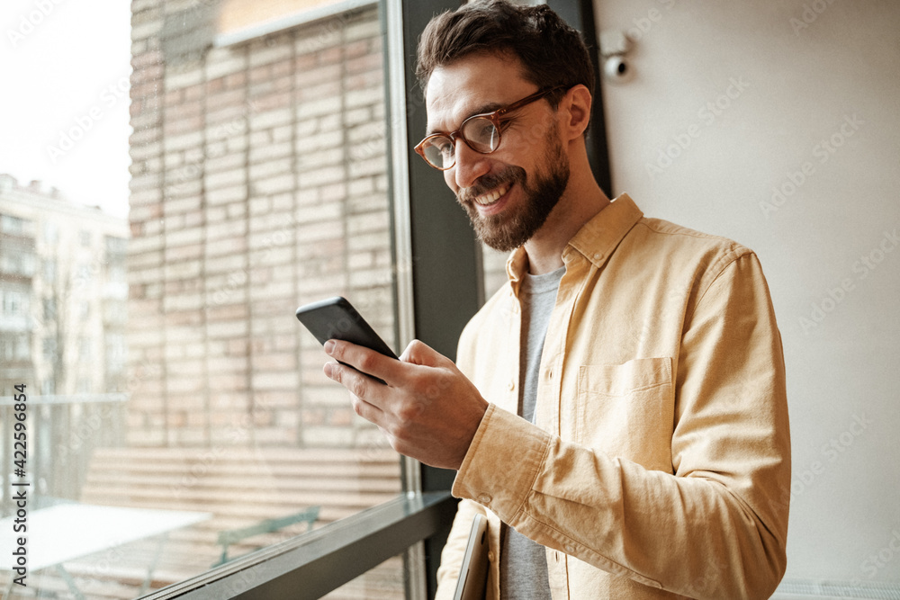 smiling and bearded man using smartphone while standing near window smiling and bearded man using smartphone while standing near window