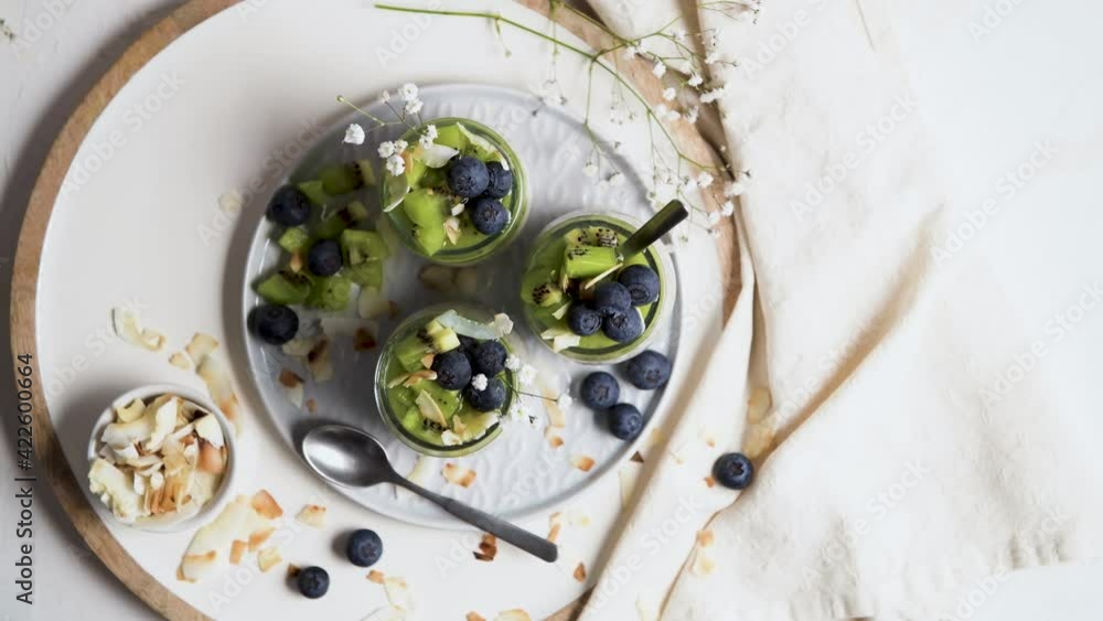 Chia pudding with kiwi, blueberries and coconut slices, three portions in glass jars on a white table.