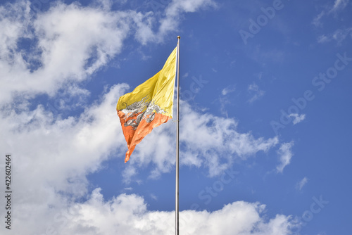 A flag of Bhutan flying with the wind on the top of a flagpole, in contrast with the blue sky and clouds.