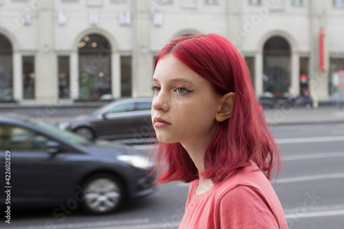 A sad lonely teenage girl with dyed red crimson hair walks along a city street.