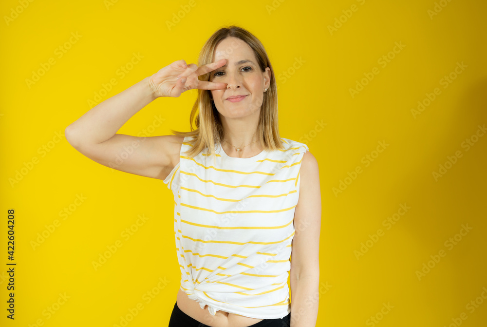 Portrait of a smiling woman showing victory sign and looking at camera isolated over yellow background Portrait of a smiling woman showing victory sign and looking at camera isolated over yellow background