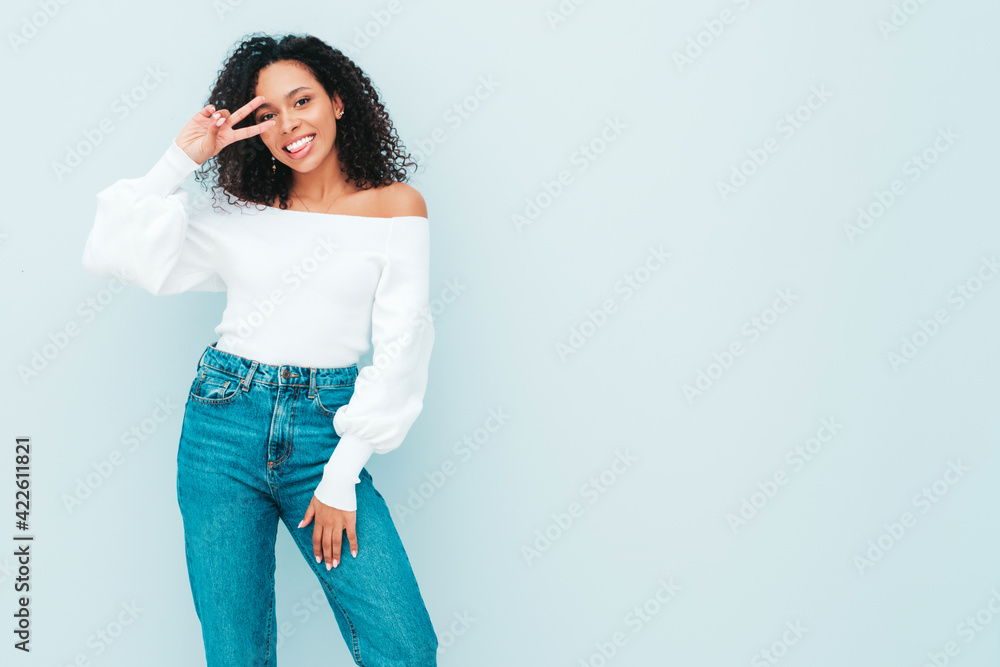 Beautiful black woman with afro curls hairstyle.Smiling model in sweater and trendy jeans clothes. Sexy carefree female posing near light blue wall in studio. Tanned and cheerful.Shows peace sign Beautiful black woman with afro curls hairstyle.Smiling model in sweater and trendy jeans clothes. Sexy carefree female posing near light blue wall in studio. Tanned and cheerful.Shows peace sign