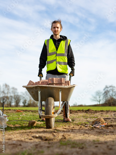 A young adult male builder wearing a high visibility vest and hard hat pushing a wheelbarrow full of bricks while on a building site