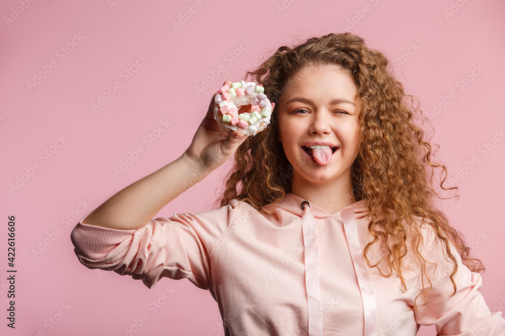 Young beautiful girl with curly hair holds a pink donut with marshmallows. Pink background. Young beautiful girl with curly hair holds a pink donut with marshmallows. Pink background.
