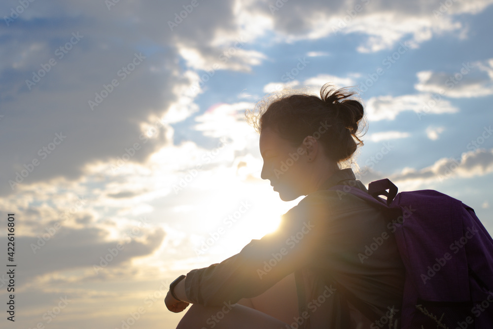 Silhouette of woman hiker with backpack sitting resting during hiking adventure. Side view of female traveler on sunset light and cloudy sky background. Overcoming difficulties to achieve the goal.
