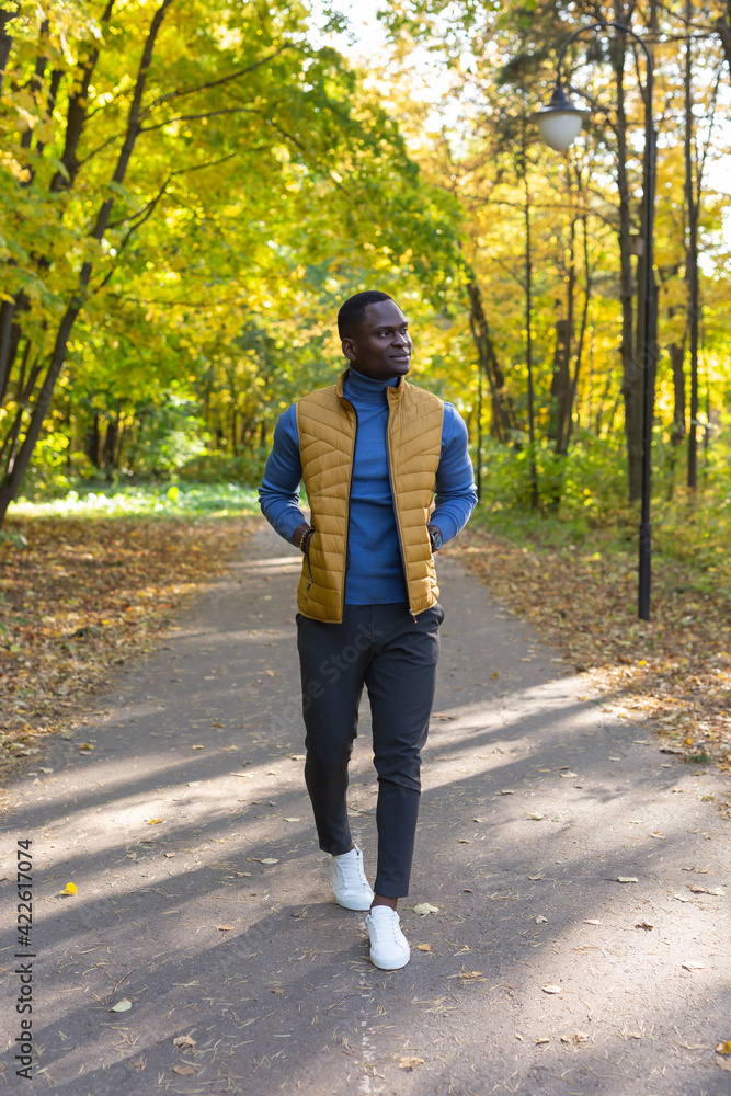 Fototapeta premium Positive african american young man in stylish clothes walks in late summer park on sunny warm warm day. Outdoor activity concept.