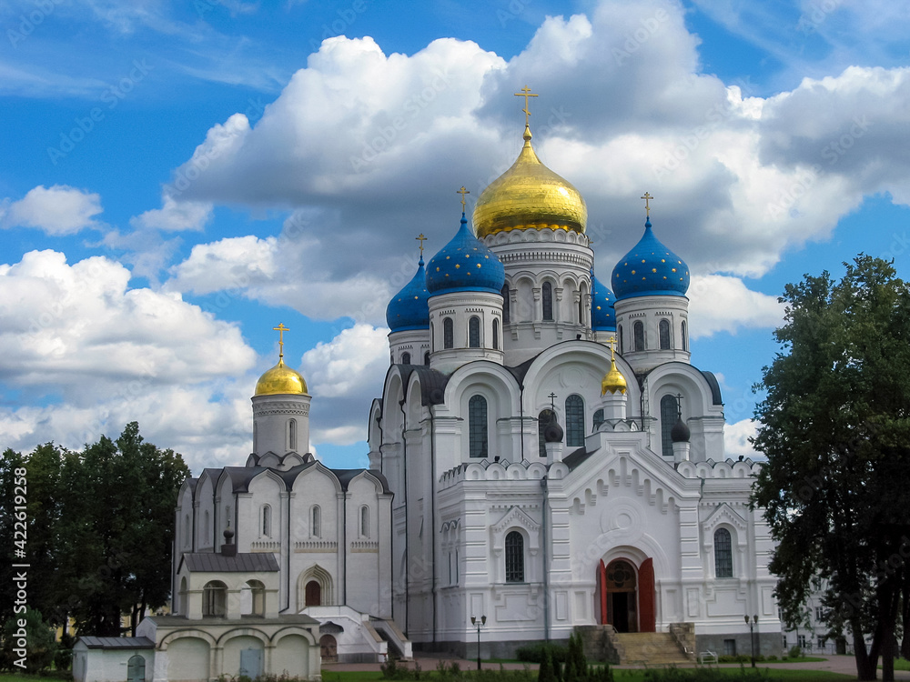 View of old Orthodox white stone church with gold and blue domes in monastery against background of green trees and cloudy sky. Religion, Christianity.