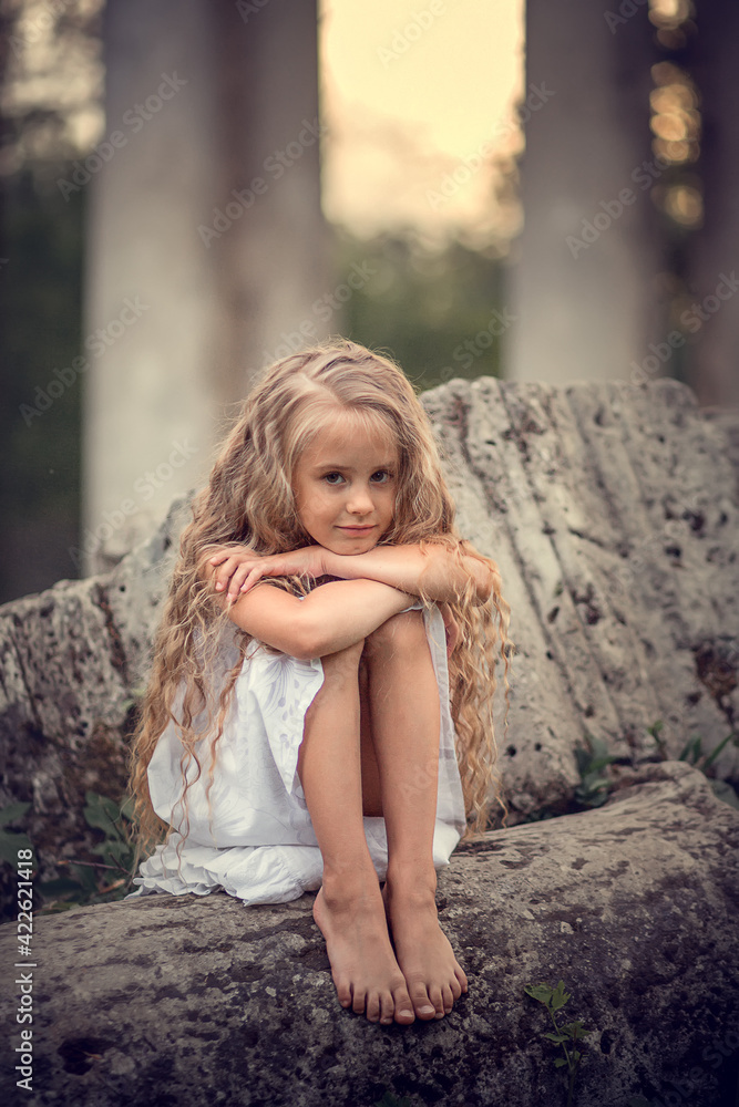 Portrait of beautiful blonde long-haired girl sitting on the ruined stones during the sunset in Pavlovsk park, Russia. Image with selective focus and toning