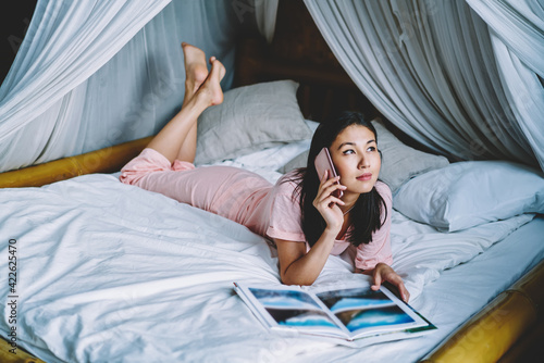 Millennial Asian woman with photo album calling to friend for discussing pictures and memoirs during weekend time in cosy home interior, attractive female in bedclothes using smartphone for talking
