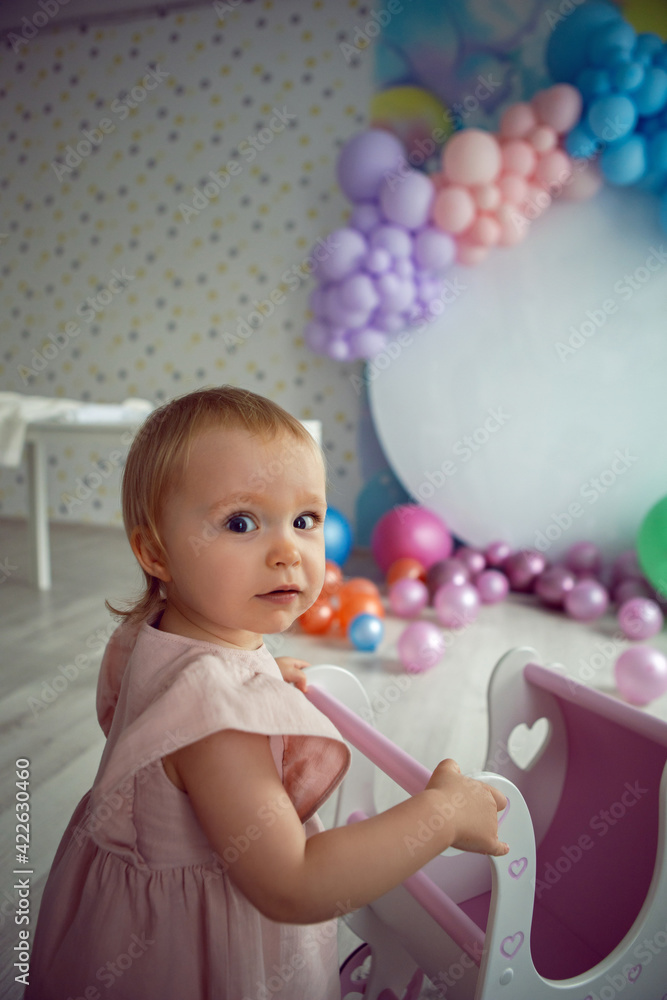 baby girl in a pink dress carries a wooden stroller toy on a background of colorful balloons and a white round background