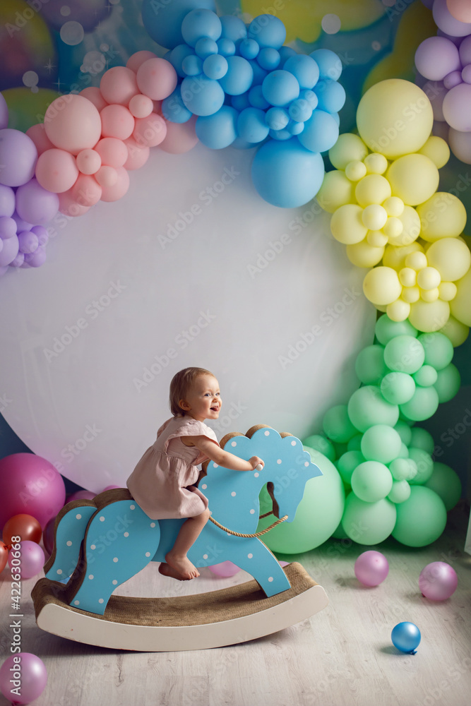 Obraz premium baby girl in a pink dress sits on a horse rocking toy on a background of colorful balloons and a white round background
