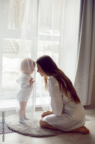 mother in a white robe sits with a child a blonde daughter at a large window of the house