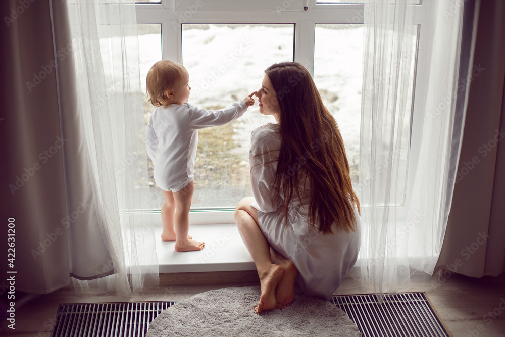 mother in a white robe sits with a child a blonde daughter at a large window of the house