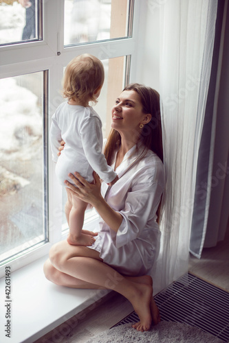 mother in a white robe sits with a child a blonde daughter at a large window of the house