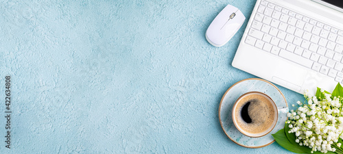Home office desk table with coffee cup, bouquet of spring flowers lily of the valley flat lay. Workplace with laptop and coffee on blue background. Blogging, freelance, florist concept. Сopy space