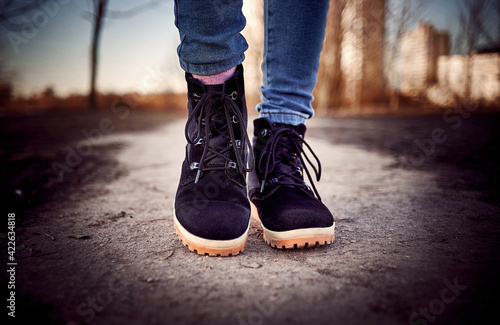 Women's feet in black leather boots walking on the small ground road. Black shoes and blue jeans.