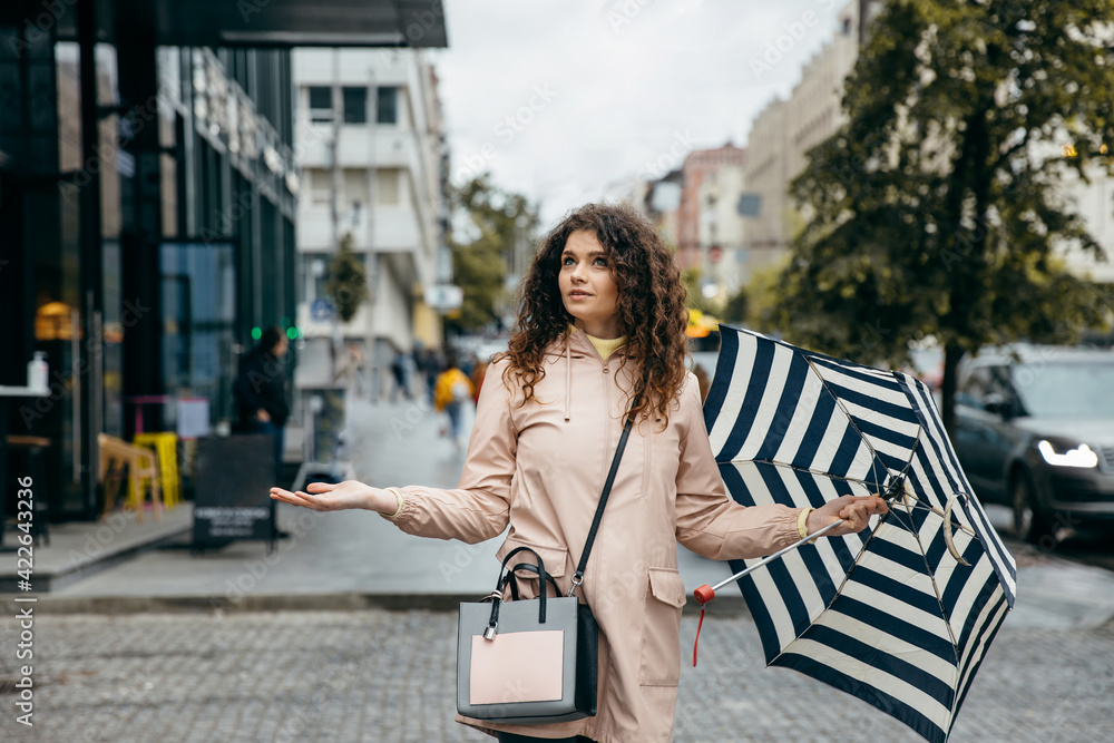Charming young curly mestizo woman under umbrella walking the street of megapolis city in rainy day