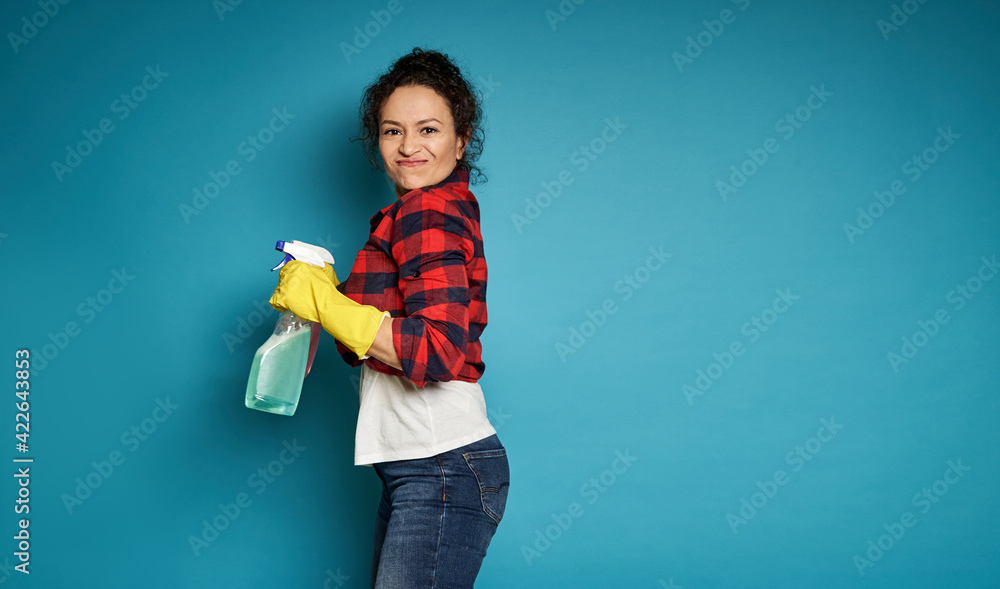 Side view to a mixed race woman holding a detergent cleaning spray and looking at the camera, posing over blue background with copy space Side view to a mixed race woman holding a detergent cleaning spray and looking at the camera, posing over blue background with copy space