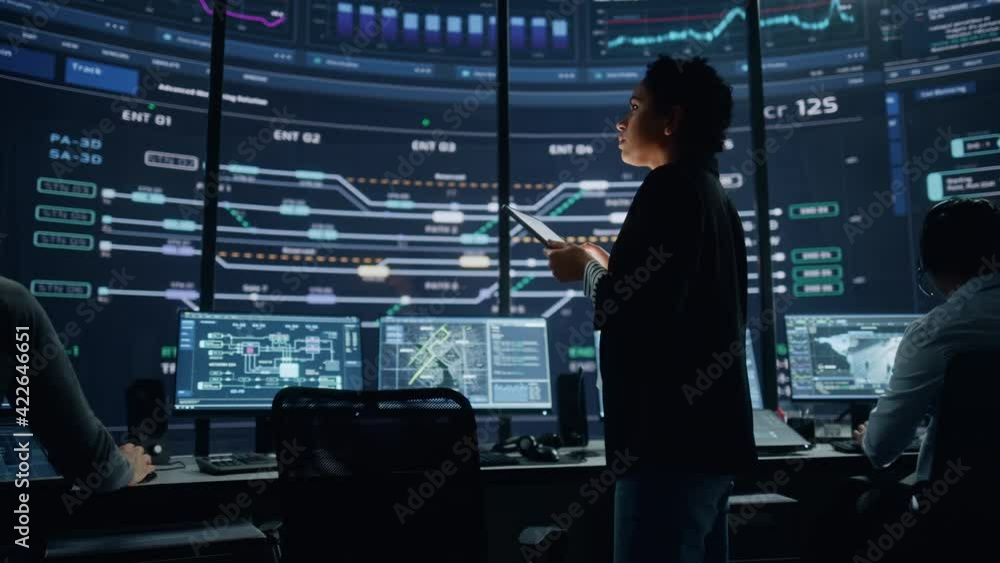 Young Multiethnic Female Government Employee Uses Tablet Computer in System Control Monitoring Center. In the Background Her Coworkers at Their Workspaces with Many Displays Showing Technical Data. Young Multiethnic Female Government Employee Uses Tablet Computer in System Control Monitoring Center. In the Background Her Coworkers at Their Workspaces with Many Displays Showing Technical Data.