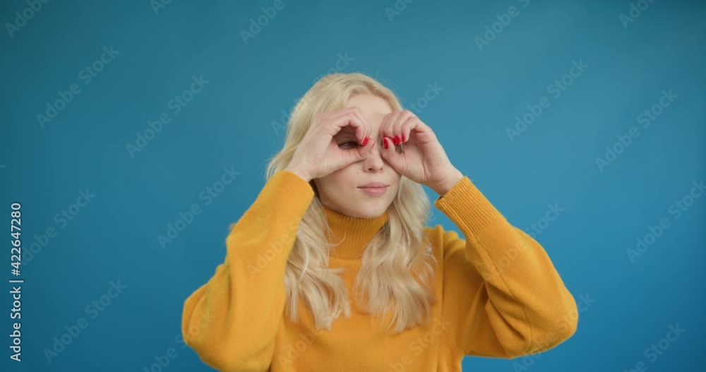 Nosy young woman making glasses shape with hands, looking through fingers in binoculars gesture and expressing surprise happy. studio shot isolated on blue background Nosy young woman making glasses shape with hands, looking through fingers in binoculars gesture and expressing surprise happy. studio shot isolated on blue background
