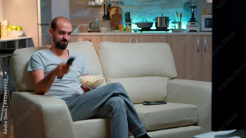 Bored man switching TV channels, holding popcorn in hads late at night while trying to watch entertainment television. Using remote control to choose favourite station and enjoy quality time Bored man switching TV channels, holding popcorn in hads late at night while trying to watch entertainment television. Using remote control to choose favourite station and enjoy quality time