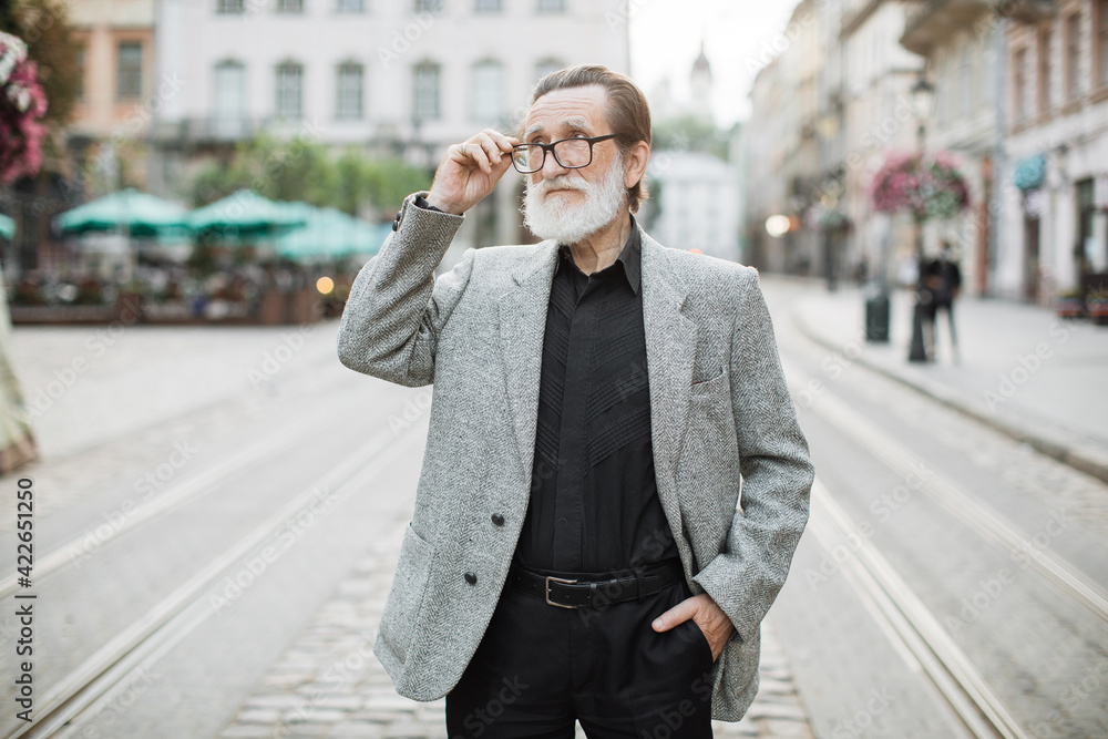 Portrait of stylish bearded man adjusting eyeglasses while standing on old city street. Positive elder male spending free time on fresh air. Blur background.