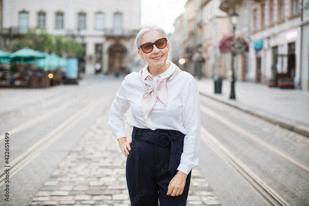 Adult grey-haired woman in elegant outfit and trendy sunglasses posing and smiling outdoors. City street on background. Happy pensioner. Adult grey-haired woman in elegant outfit and trendy sunglasses posing and smiling outdoors. City street on background. Happy pensioner.