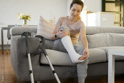 Woman in her late twenties on couch at home with elbow crutches and orthopedic plaster. Fracture of the leg or foot. Concept of rehabilitation and healing physiotherapy. Orthopedics and Traumatology.