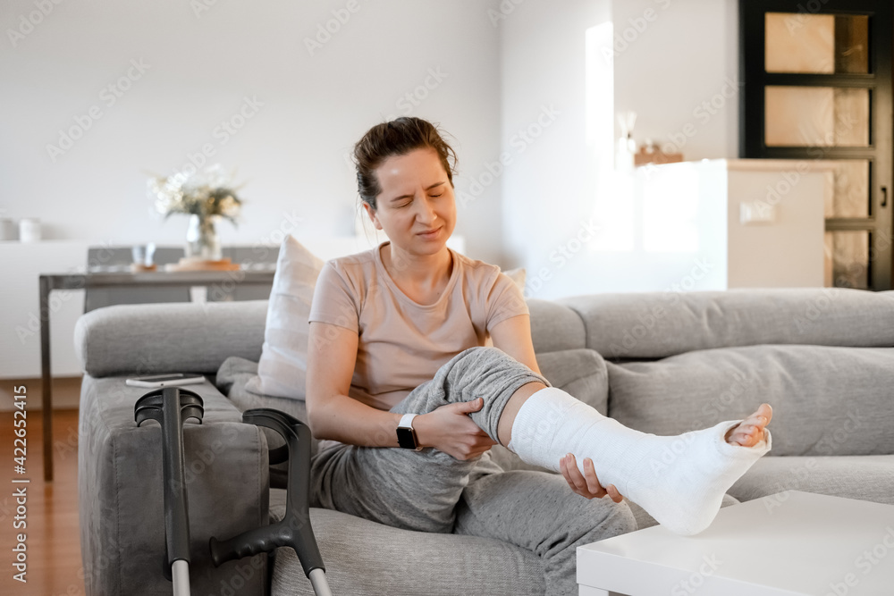 Young woman with pain expression on couch at home with orthopedic plaster massaging leg. Fracture of the leg or foot. Concept of rehabilitation and healing physiotherapy. Orthopedics and Traumatology. Young woman with pain expression on couch at home with orthopedic plaster massaging leg. Fracture of the leg or foot. Concept of rehabilitation and healing physiotherapy. Orthopedics and Traumatology.