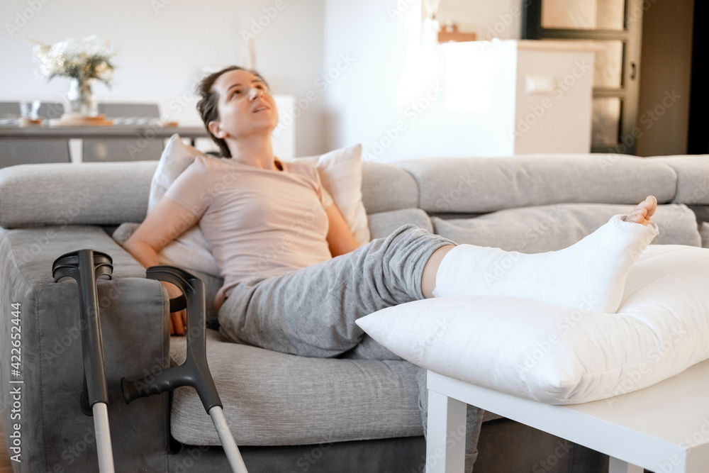Young woman with pain expression on couch at home with orthopedic plaster massaging leg. Fracture of the leg or foot. Concept of rehabilitation and healing physiotherapy. Orthopedics and Traumatology. Young woman with pain expression on couch at home with orthopedic plaster massaging leg. Fracture of the leg or foot. Concept of rehabilitation and healing physiotherapy. Orthopedics and Traumatology.