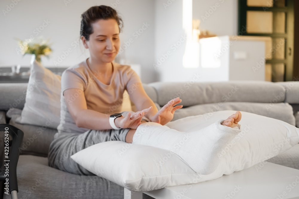Woman in her late twenties on couch at home with orthopedic plaster massaging leg. Fracture of the leg or foot. Concept of rehabilitation and healing physiotherapy. Orthopedics and Traumatology. Woman in her late twenties on couch at home with orthopedic plaster massaging leg. Fracture of the leg or foot. Concept of rehabilitation and healing physiotherapy. Orthopedics and Traumatology.