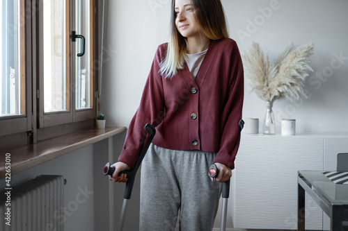 Adult woman in her late twenties on crutches at home is looking into the window with hope. Fracture of the leg or foot. Concept of rehabilitation and healing. Orthopedics and Traumatology photo