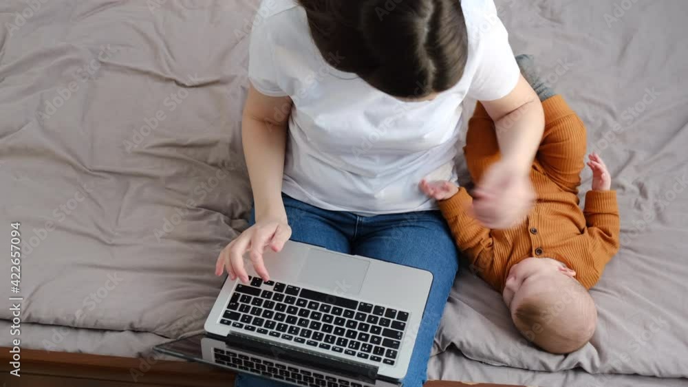 Top view of young business mother using laptop, sitting on bed near sweet baby girl at home, beautiful caring mom shopping or chatting online in social network, freelancer working on computer project Top view of young business mother using laptop, sitting on bed near sweet baby girl at home, beautiful caring mom shopping or chatting online in social network, freelancer working on computer project