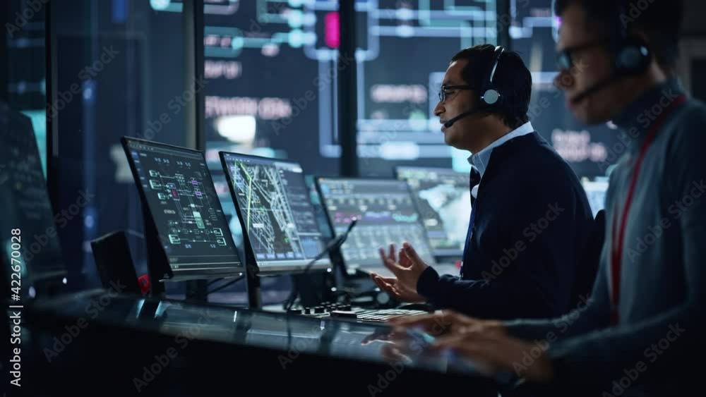 Portrait of Professional IT Technical Support Specialist Working on Computer in Monitoring Control Room with Digital Screens. Employee Wears Headphones with Mic and Talking on a Call. Portrait of Professional IT Technical Support Specialist Working on Computer in Monitoring Control Room with Digital Screens. Employee Wears Headphones with Mic and Talking on a Call.