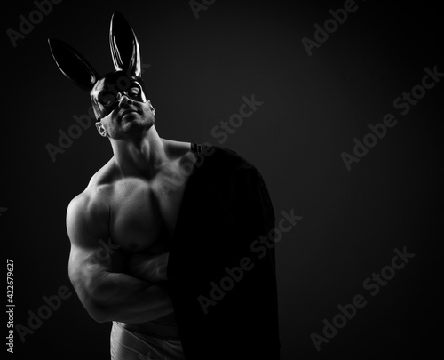Naked topless muscular man in underwear, black rabbit mask and with jacket on shoulder stands at copy space over dark background