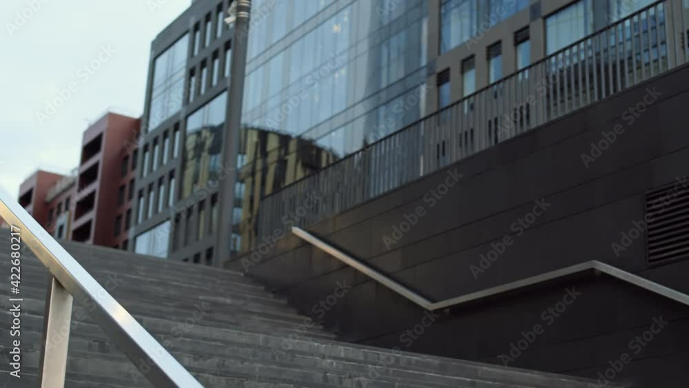 Arc of childish brown-haired Caucasian woman smiling, wearing summer dress, holding paper shopping bags in hands, sliding down metal handrail of stairs outside modern buildings Arc of childish brown-haired Caucasian woman smiling, wearing summer dress, holding paper shopping bags in hands, sliding down metal handrail of stairs outside modern buildings
