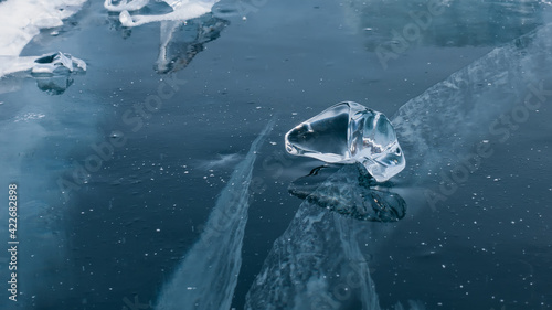 Fototapeta Naklejka Na Ścianę i Meble -  A transparent piece of ice of a bizarre shape lies on the thick blue ice with cracks. Mirror reflection. Lake Baikal