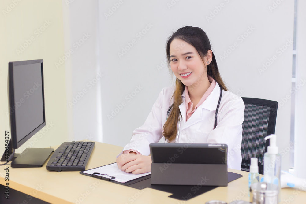 Asian young woman doctor in a white lab coat is sitting on office room at hospital while has a computer, clipbroad and alcohol gel on the deck. Asian young woman doctor in a white lab coat is sitting on office room at hospital while has a computer, clipbroad and alcohol gel on the deck.