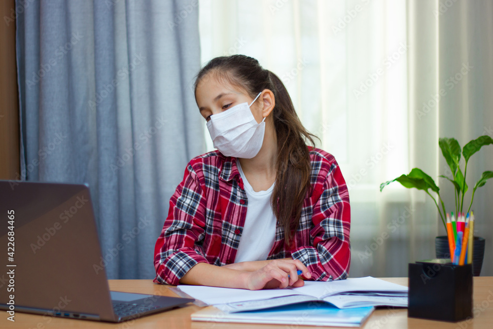 Bored brunette girl, dressed in a medical mask, red shirt, sits at home studying, writes homework, communicates on the Internet at the computer. 