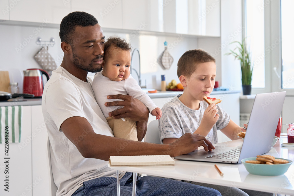 Young Black father, African dad remote working or distance learning online from home office using laptop computer having breakfast holding mixed race baby sitting at kitchen table with kid son at home Young Black father, African dad remote working or distance learning online from home office using laptop computer having breakfast holding mixed race baby sitting at kitchen table with kid son at home