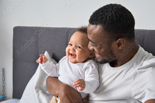 Happy loving young Black dad holding adorable mixed race baby daughter having fun in bed at home. Smiling African father playing with cute funny infant child girl waking up in bedroom in the morning.