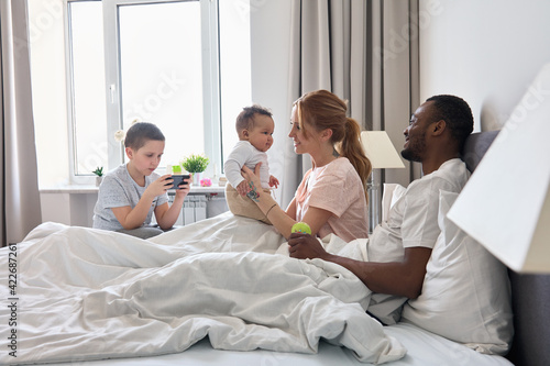 Happy multiracial family with kids having fun sitting in bed together. Mixed race young parents African father and Caucasian mother holding baby daughter while teen son using phone at home.