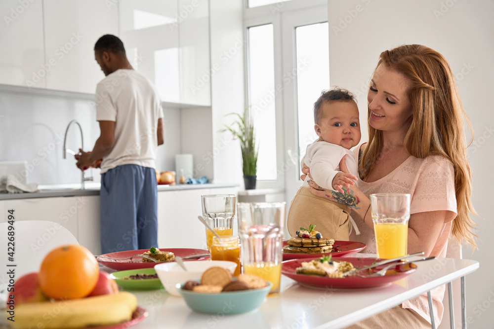 Happy multiethnic family with child having breakfast in kitchen in the morning. Caucasian mom holding cute adorable funny infant mixed race baby daughter playing while African dad making meal at home. Happy multiethnic family with child having breakfast in kitchen in the morning. Caucasian mom holding cute adorable funny infant mixed race baby daughter playing while African dad making meal at home.